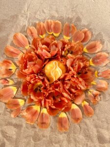 Orange flowers are arranged in a Yoni Mandala formation on the sand.