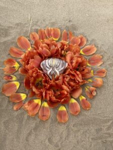 A Yoni Mandala is created with an orange flower placed in the sand.