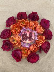 A Yoni Mandala with a pomegranate and orange flower arrangement on the sand.